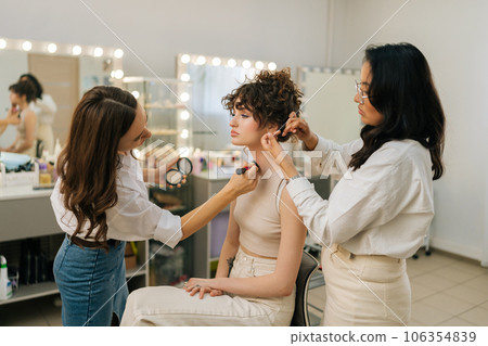 Side view of professional makeup artist applying foundation brush on neck body of young woman model. Hairdresser doing stylish hairdo to curly haired female. Concept of four-handed makeup and styling. Side view of professional makeup artist applying foundation brush on neck body of young woman model. Hairdresser doing stylish hairdo to curly haired female. Concept of four-handed makeup and styling. 106354839