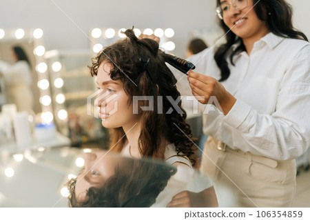 Smiling female hairstylist using hair iron and comb for young woman hairstyling in modern hairdressing studio. Hairdresser creating female hairdo with tongs. Concept of fashion and beauty. 106354859