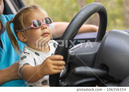 A cute little girl driving a car on her father's lap. Child girl in sunglasses with a serious expression learns to steer a car. A father teaches his daughter how to drive a car. 106356354