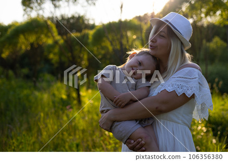 The daughter gently clings to her mother sitting in her arms. In summer, against the backdrop of the setting sun and nature, a mother lovingly holds a small child in her arms. The daughter gently clings to her mother sitting in her arms. In summer, against the backdrop of the setting sun and nature, a mother lovingly holds a small child in her arms. 106356380