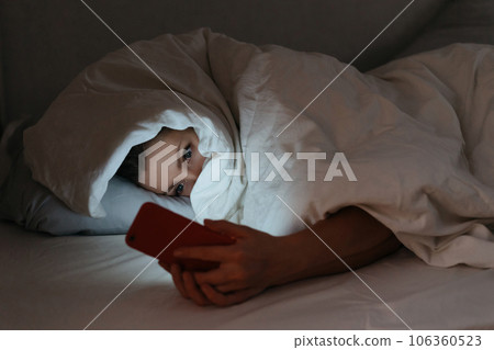 Young woman laying in bed at night while staring at her cellphone. Young woman laying in bed at night while staring at her cellphone. 106360523