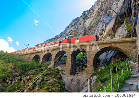 Red train passing by Devil's bridge at St. Gotthard pass on the Swiss Alps in Andermatt, Switzerland Red train passing by Devil's bridge at St. Gotthard pass on the Swiss Alps in Andermatt, Switzerland 106361684
