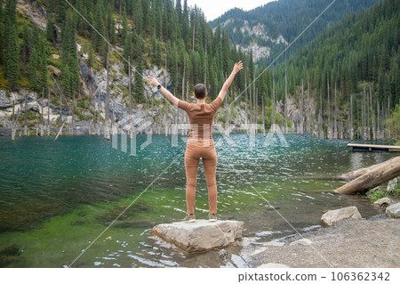 Tourist woman travels through the mountains of Central Asia, standing with arms outstretched and looking at the mountain lake Kaindy with sunken fir trees Tourist woman travels through the mountains of Central Asia, standing with arms outstretched and looking at the mountain lake Kaindy with sunken fir trees 106362342