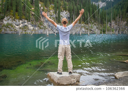 Tourist man travels through the mountains of Central Asia, standing with arms outstretched and looking at the mountain lake Kaindy with sunken fir trees Tourist man travels through the mountains of Central Asia, standing with arms outstretched and looking at the mountain lake Kaindy with sunken fir trees 106362366