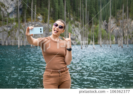 A young woman travels through the Tien Shan mountains of Central Asia, makes a cheerful selfie against the backdrop of the picturesque mountain lake Kaindy in the mountains of Kazakhstan A young woman travels through the Tien Shan mountains of Central Asia, makes a cheerful selfie against the backdrop of the picturesque mountain lake Kaindy in the mountains of Kazakhstan 106362377