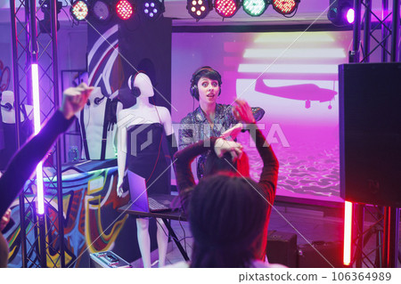 Young woman dj performing on stage and engaging people to party at nightclub discotheque. Excited girl in headphones playing electronic music while crowd clubbing on dancefloor Young woman dj performing on stage and engaging people to party at nightclub discotheque. Excited girl in headphones playing electronic music while crowd clubbing on dancefloor 106364989
