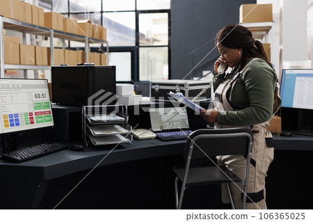 African american employee holding clipboard, analyzing goods inventory report in storehouse. Storage room supervisor talking at landline phone with remote client discussing delivery details 106365025