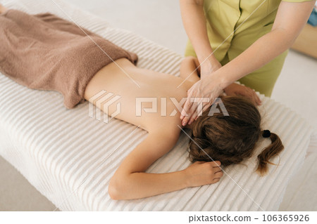 Closeup top view of pretty relaxed girl having neck, shoulder and back massage by unrecognizable female masseuse lying on massage table. Adorable kid getting physiotherapy from masseuse for body care 106365926