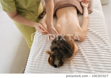 High-angle view of unrecognizable female pediatric masseuse doing therapeutic massage to relax shoulder joint and shoulder blade of little girl. Kid client get service massage in spa salon. 106365927