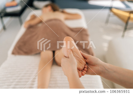 Close-up hands of unrecognizable female masseuse doing foot and legs massage to cute ten-year-old girl lying on massage table in spa salon. Masseur kneads foot and leg of little kid in clinic. Close-up hands of unrecognizable female masseuse doing foot and legs massage to cute ten-year-old girl lying on massage table in spa salon. Masseur kneads foot and leg of little kid in clinic. 106365931