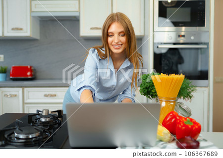 Young blonde woman with long hair using laptop in a kitchen Young blonde woman with long hair using laptop in a kitchen 106367689