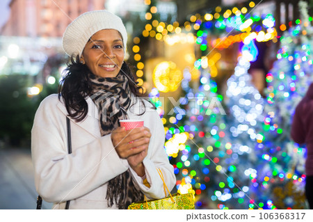 Cheerful Latina with cup of hot tea walking at Christmas street fair Cheerful Latina with cup of hot tea walking at Christmas street fair 106368317