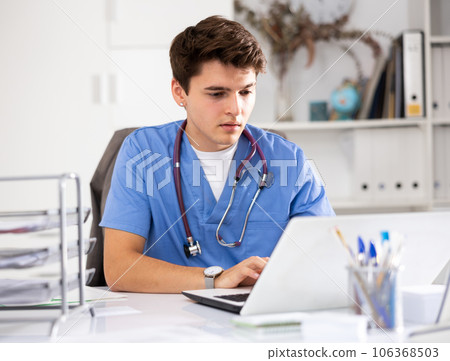 Portrait of man doctor in surgical scrubs sitting at working table in office and using laptop 106368503