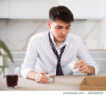 Sick young man with hangover drinks pills while sitting at table 106368505
