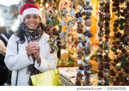 Cheerful hispanic woman in Santa hat with cup of hot tea walking at Christmas street fair Cheerful hispanic woman in Santa hat with cup of hot tea walking at Christmas street fair 106368508