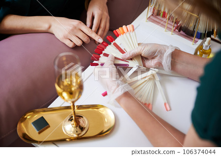 Close up of female hand pointing at red color nail sample while choosing manicure with manicurist. Manicure master in sterile gloves holding nail polish color palette. 106374044