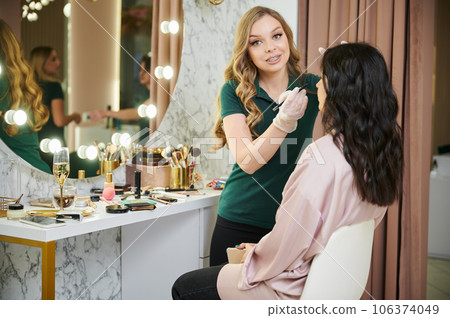 Female makeup artist applying pencil eyeliner to client upper eyelid. Beauty specialist in sterile gloves doing professional eye makeup on woman in beauty salon. 106374049