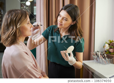Young woman beauty specialist in sterile gloves applying powder on client face with cosmetic brush. Professional makeup artist doing makeup for woman in beauty salon. Young woman beauty specialist in sterile gloves applying powder on client face with cosmetic brush. Professional makeup artist doing makeup for woman in beauty salon. 106374050
