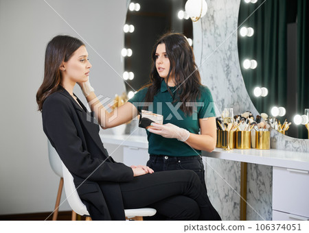 Stylish woman sitting on stool at dressing table while beauty specialist doing professional makeup in modern salon. Female beauty specialist applying cosmetics on woman face in visage studio. 106374051