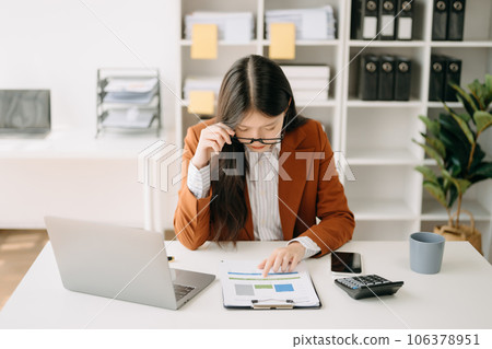Confident beautiful Asian businesswoman typing laptop computer and digital tablet while holding coffee at office 106378951