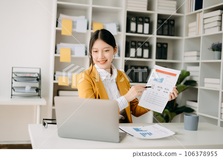 Young attractive Asian woman smiling thinking planning writing in notebook, tablet and laptop working from home, looking at camera at office Young attractive Asian woman smiling thinking planning writing in notebook, tablet and laptop working from home, looking at camera at office 106378955
