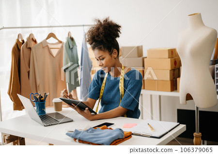 Calm curly brunette dark skinned woman on desk in modern office of fashion designer and holds tablet and smartphone. 106379065