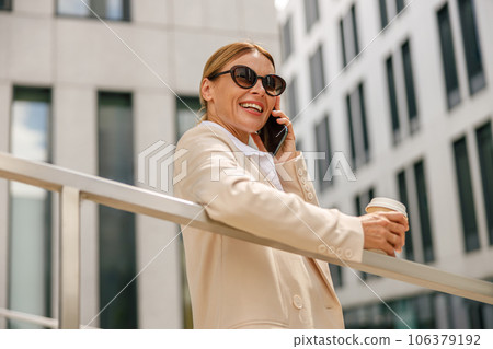 Stylish woman entrepreneur talking phone whiile standing on modern building background with coffee 106379192