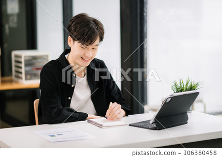Young attractive Asian man smiling thinking planning writing in notebook, tablet and laptop working from home, looking at camera at office . 106380458