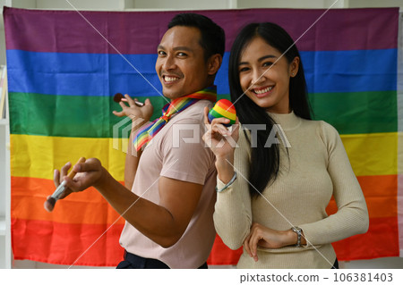 Confident gay male beauty blogger and friend standing against LGBTQ rainbow flag and smiling to camera 106381403