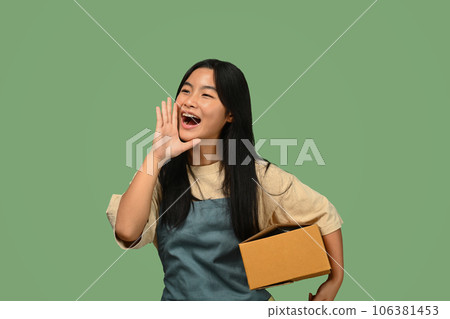 Teenage Asian girl holding parcel box and shouting with hands on mouth, standing against green background 106381453