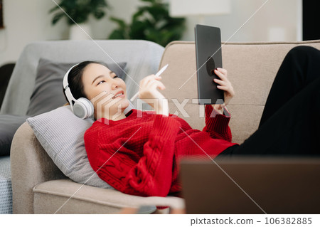 Smiling girl relaxing at home, she is playing music using smartphone tablet, laptop, and wearing white headphones. Smiling girl relaxing at home, she is playing music using smartphone tablet, laptop, and wearing white headphones. 106382885