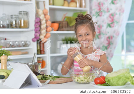 little girl preparing fresh salad on kitchen table with tablet at home little girl preparing fresh salad on kitchen table with tablet at home 106383592