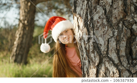 A young woman in a red Christmas hat looks out from behind a big tree and smiles. Christmas in a warm forest. A young woman in a red Christmas hat looks out from behind a big tree and smiles. Christmas in a warm forest. 106387930