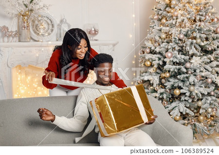 Young black couple with gifts in Christmas eve. Man sitting on a coach at home. Brunette woman standing behind and presented a gift for her boyfriend. 106389524