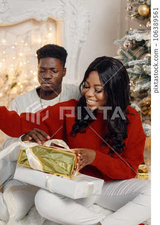 Happy black couple holding gifts and sitting near Christmas tree. Man amd woman celebrating winter holidays at home. Brunette woman wearing a red sweater. Happy black couple holding gifts and sitting near Christmas tree. Man amd woman celebrating winter holidays at home. Brunette woman wearing a red sweater. 106389561