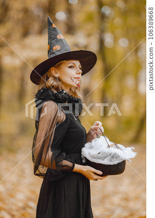 Blonde young girl witch in forest on Halloween. Girl wearing black dress and cone hat. Witch holding a magician stuff. Blonde young girl witch in forest on Halloween. Girl wearing black dress and cone hat. Witch holding a magician stuff. 106389668