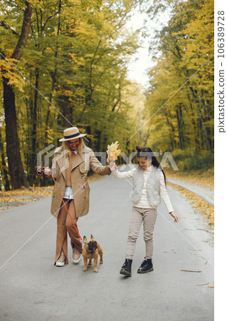 Woman, little girl and dog walking in the autumn park. Brown french bulldog with female owners spend a day at the park playing and having fun. Woman wearing beige coat and a hat. 106389728