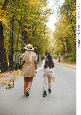Woman, little girl and dog walking in the autumn park. Brown french bulldog with female owners spend a day at the park playing and having fun. Woman wearing beige coat and a hat. 106389730