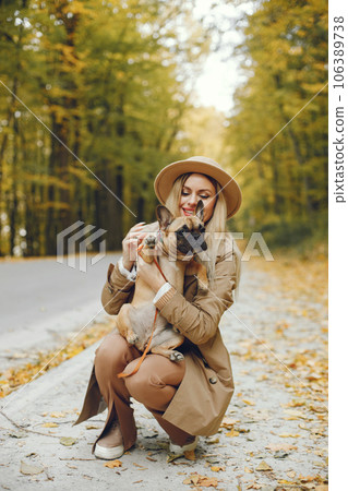 Woman and dog play and have fun in the autumn park. Brown french bulldog with female owner spend a day at the park walking. Woman wearing beige coat and a hat. 106389738