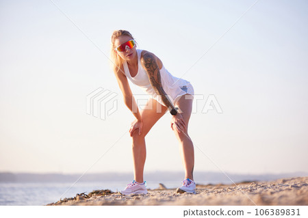 Tired fit woman dressed sporty after running, jogging hard outdoors on beach in morning over sunrise. Everyday fitness routine. 106389831