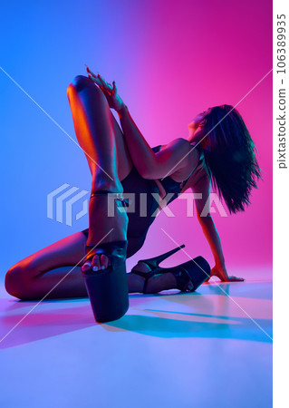 Beautiful young woman wearing sexy high heels and black bodysuit while dancing in dance studio in blue-pink neon light. 106389935