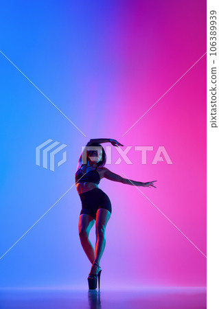 Beautiful female dancer wearing black and high heels while performing pole dance tricks in gradient neon light 106389939