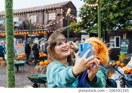 Two laughing female friends in fun glasses and witch hat having fun and making selfie on phone on decorated pumpkin farm. Selecting Thanksgiving and Halloween holidays decor. Autumn fall festive mood. 106391395