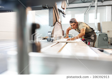 Young carpenter cutting a piece of wood in using a circular saw in furniture factory 106392913