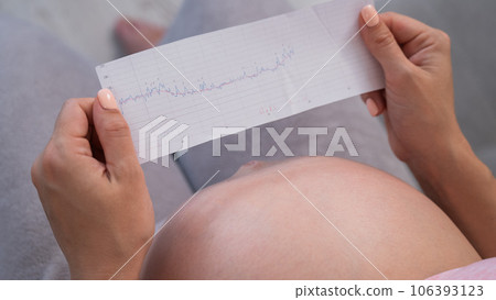 A pregnant woman looks at the results of cardiotocography while sitting on the couch. Close up of the belly. 106393123