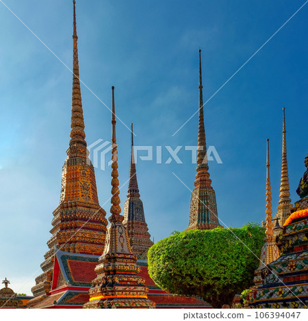 A Thai temple complex with ornate chedis decorated with gold and colorful tiles under a clear blue sky. 106394047