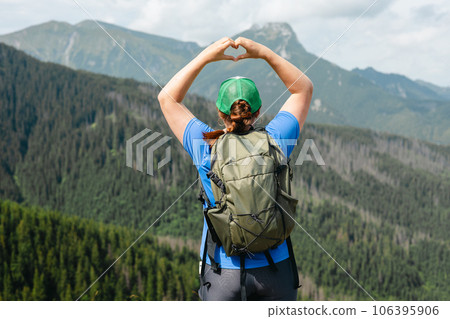 Woman showing heart shape on nature green background. Happy Girl wearing green hat with backpack. Freedom, happiness, travel and vacations concept, outdoor activities 106395906