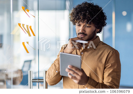 Serious thinking businessman standing near a glass board with colored papers and stickers, hispanic man holding a tablet computer in his hands, thinking about financial strategy and economic plans. Serious thinking businessman standing near a glass board with colored papers and stickers, hispanic man holding a tablet computer in his hands, thinking about financial strategy and economic plans. 106396079