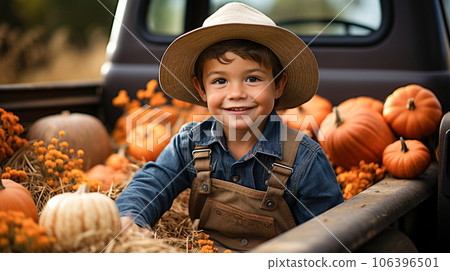 Delighted smiling young boy wearing overalls and a cowboy hat sitting in the back of a truck filled with hay and pumpkins - generative AI. 106396501