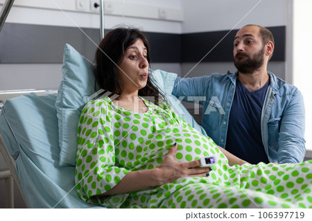 Pregnant woman doing breathing exercices, holding hands on belly while having painful contractions. Patient with pregnancy going into labor in hospital ward, husband standing beside her comforting. 106397719